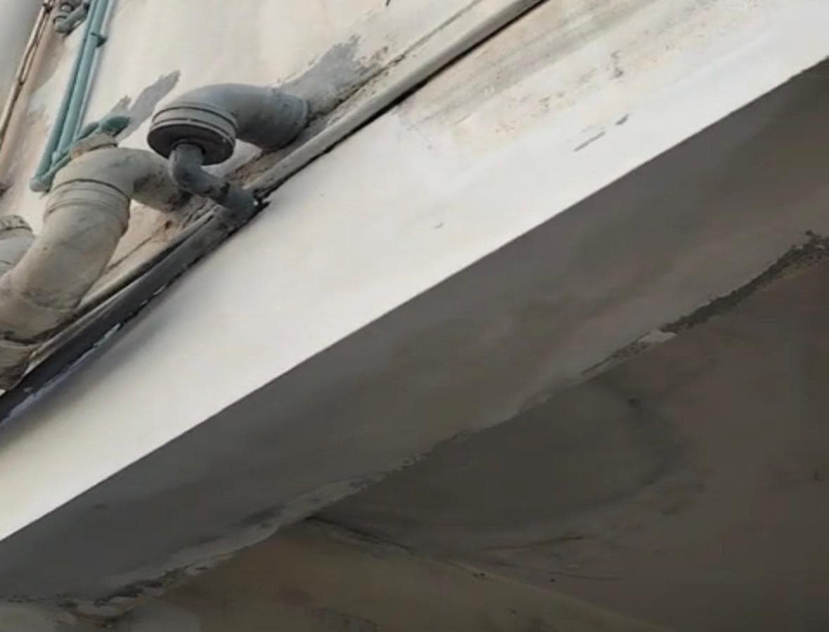 A repaired concrete soffit and parapet junction on a Zinj building, with rainwater downpipes passing through the slab edge and a fresh sealed joint running along it